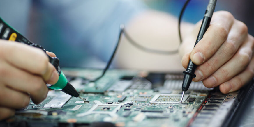 Closeup shot of male hands testing electric current voltage in circuit board of disassembled laptop using multimeter tool on table in maintenance shop